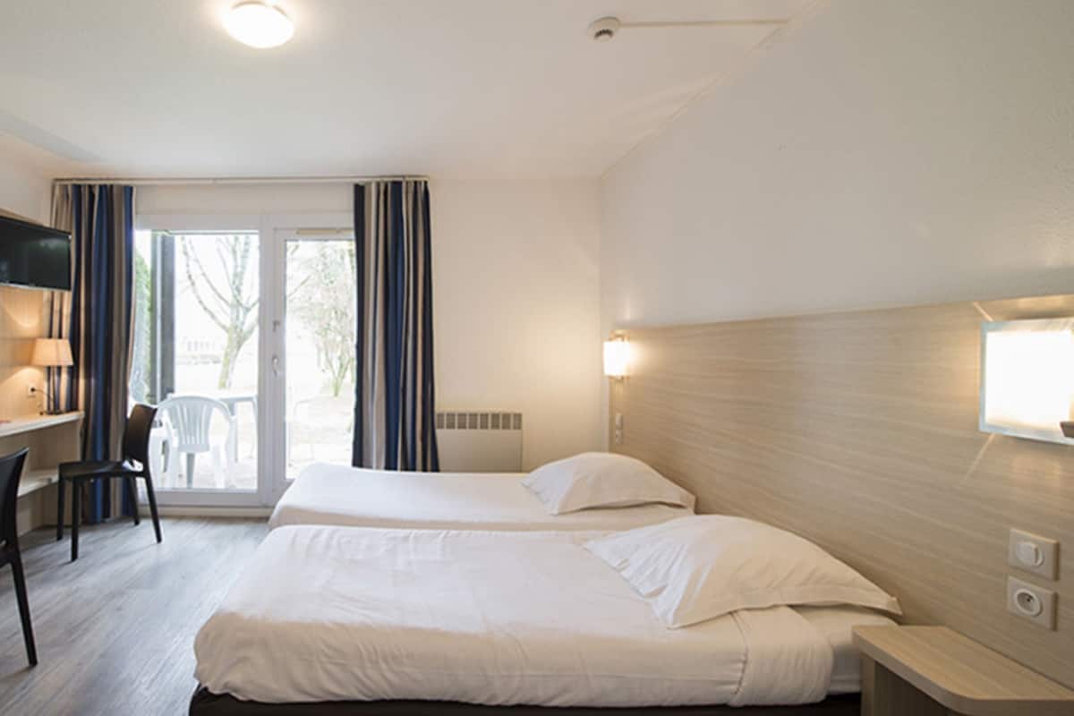 A hotel room with two beds, a desk, a chair, and a sliding glass door leading to an outdoor area.