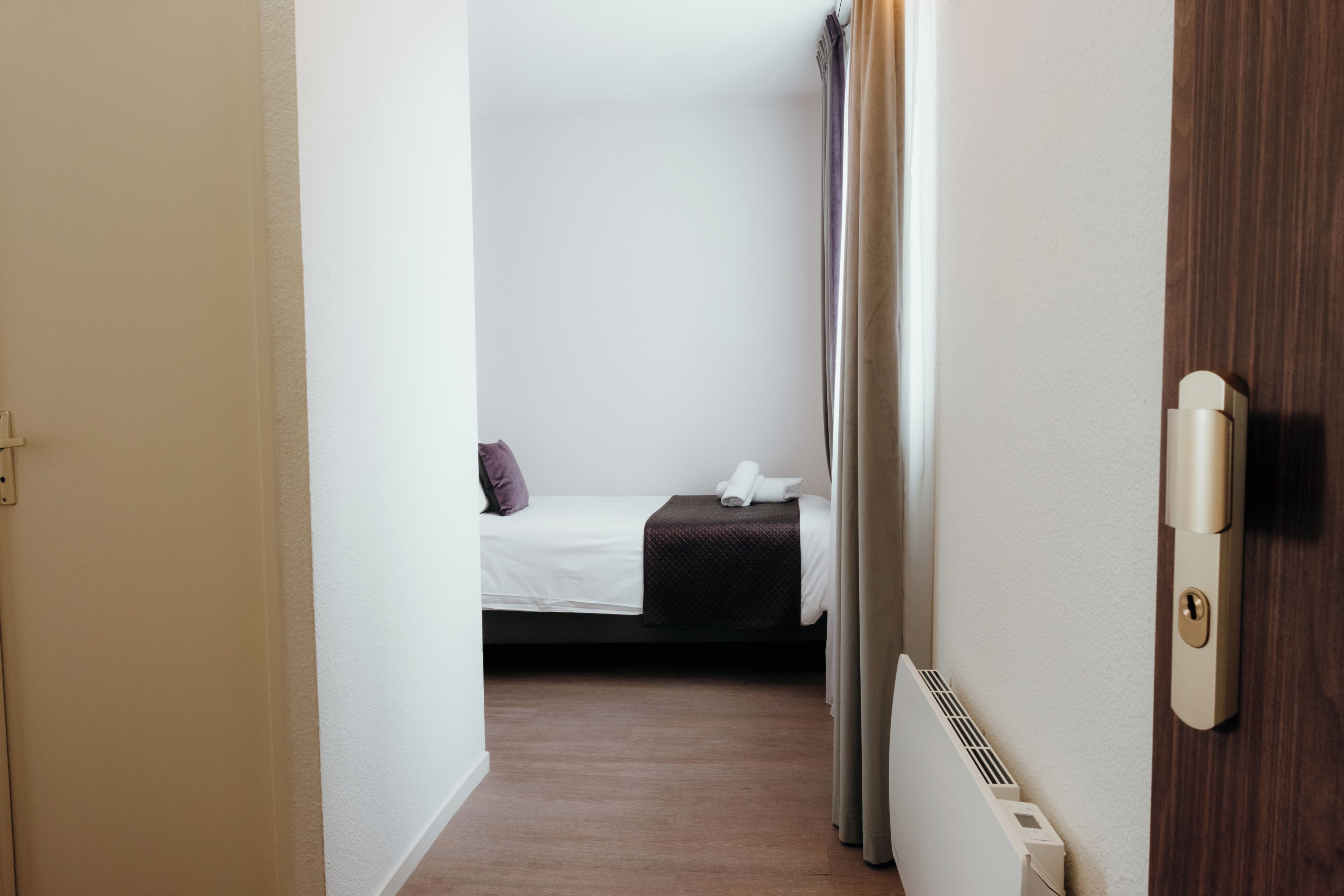 Een smalle gang die leidt naar een kleine slaapkamer met een bed, een kussen en een opgerolde handdoek.