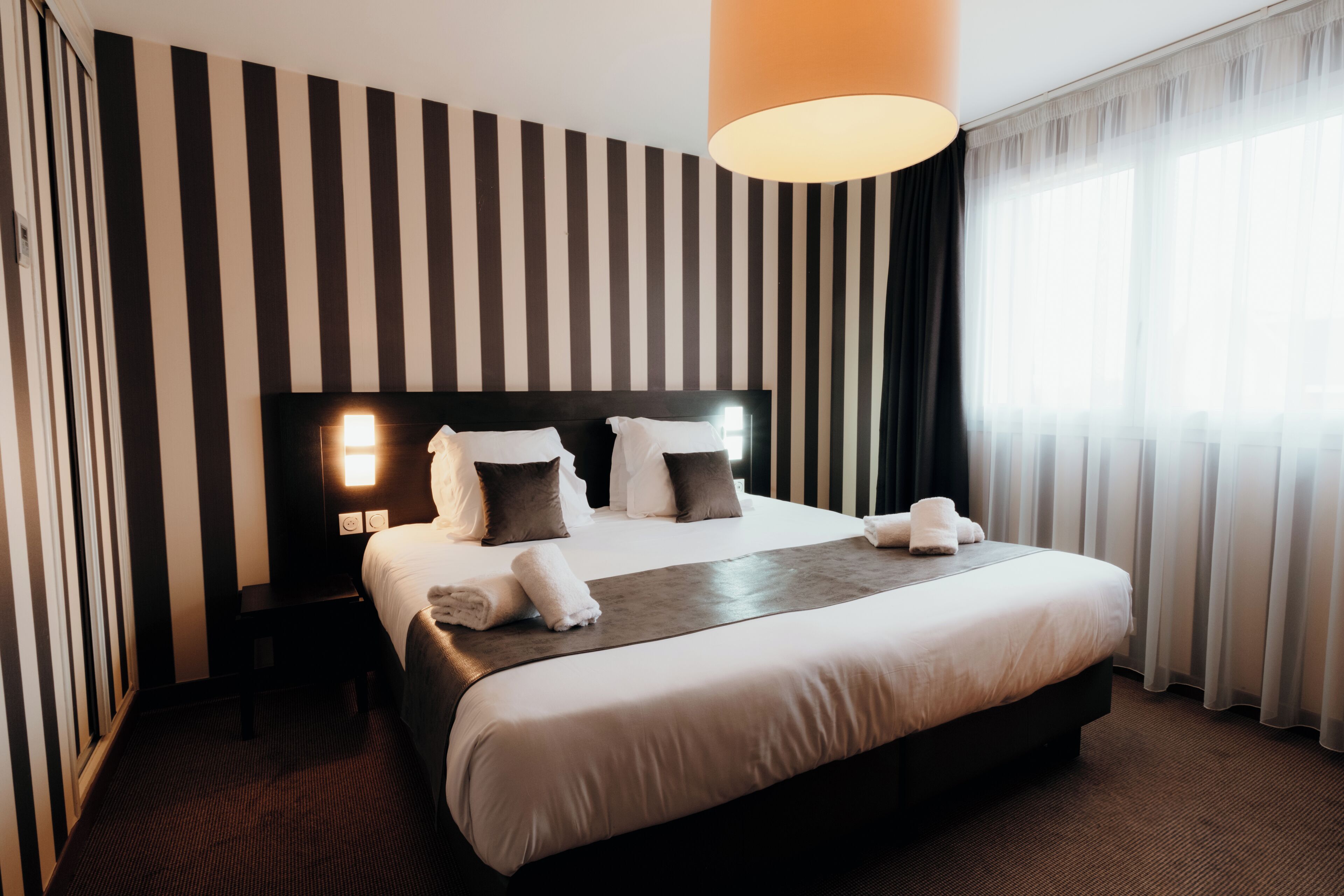 A hotel room with a large bed, a headboard with a light, a bedside table, and a window with curtains.