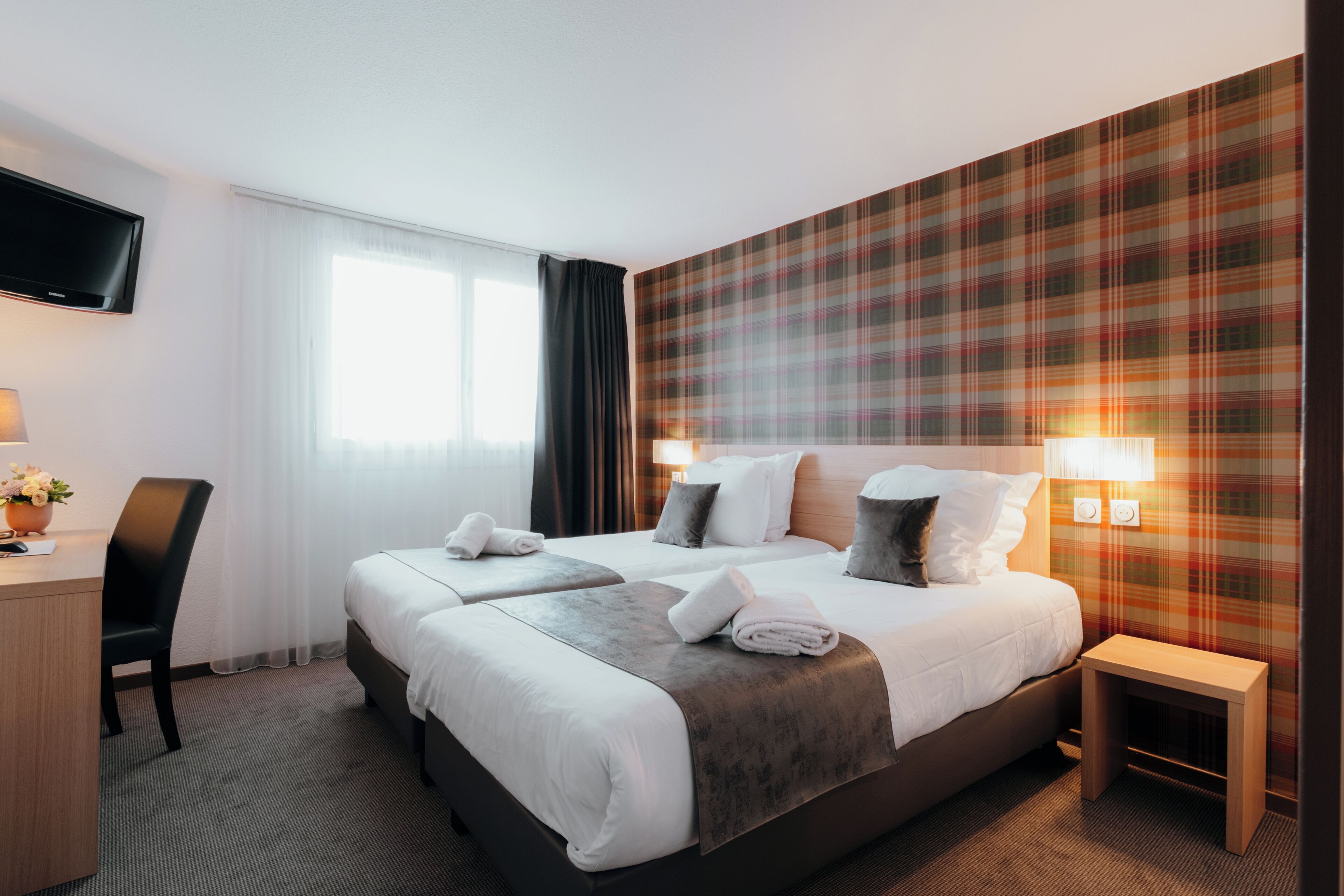 A hotel room with two beds, a desk, a chair, a TV, and a plaid-patterned wall.