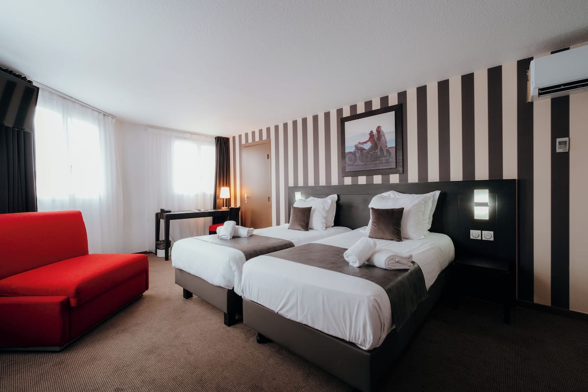 A hotel room with two beds, a red sofa, a desk, and a window with curtains.