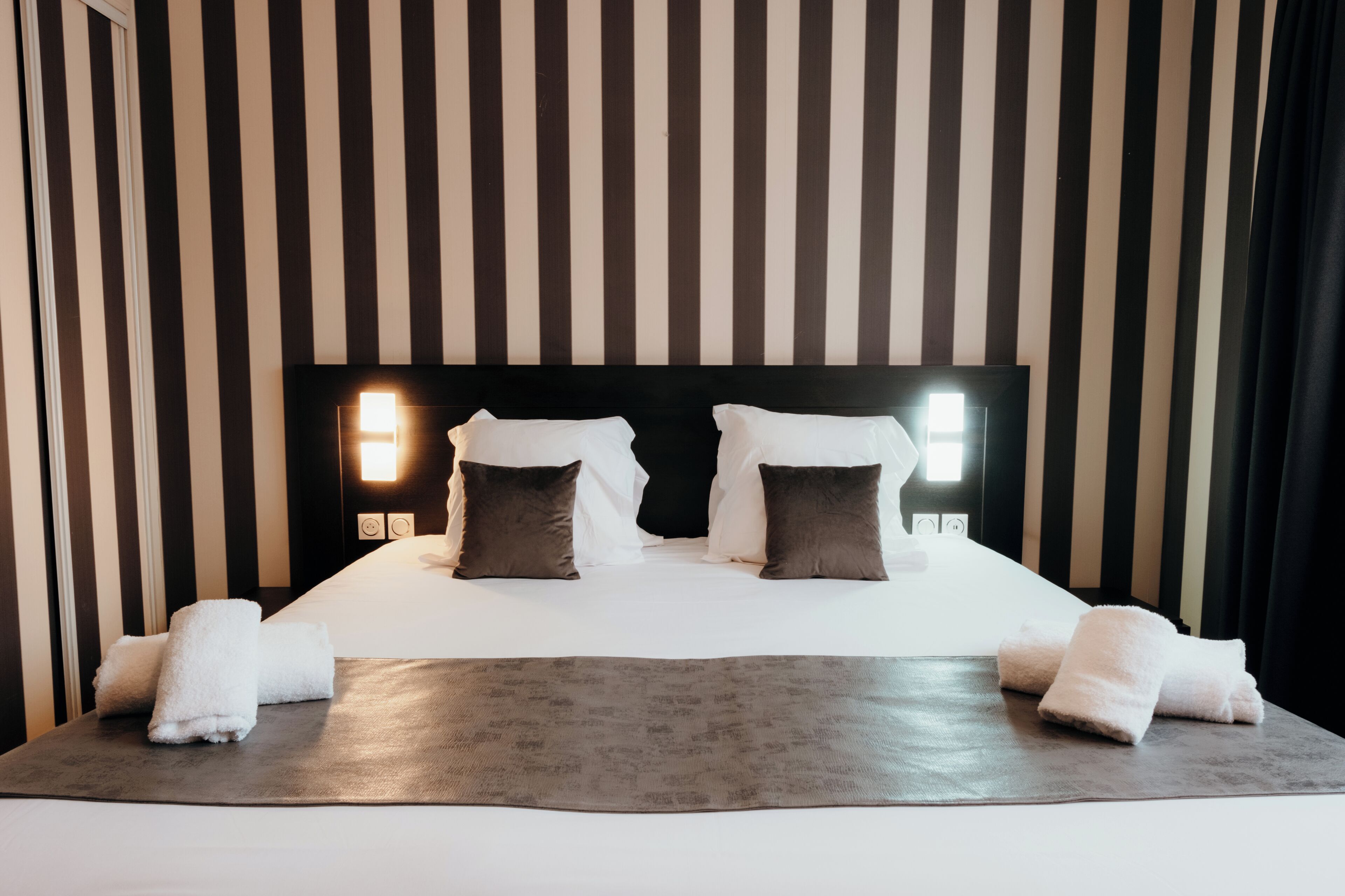 Un lit bien fait, avec du linge de lit blanc et deux oreillers, une couverture grise et deux serviettes blanches posées sur un cadre de lit en bois foncé.