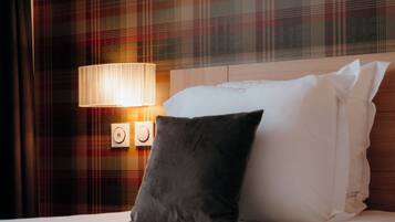 A hotel room with a plaid-patterned wall, a bedside lamp, a bed with white linens, and a dark gray pillow.