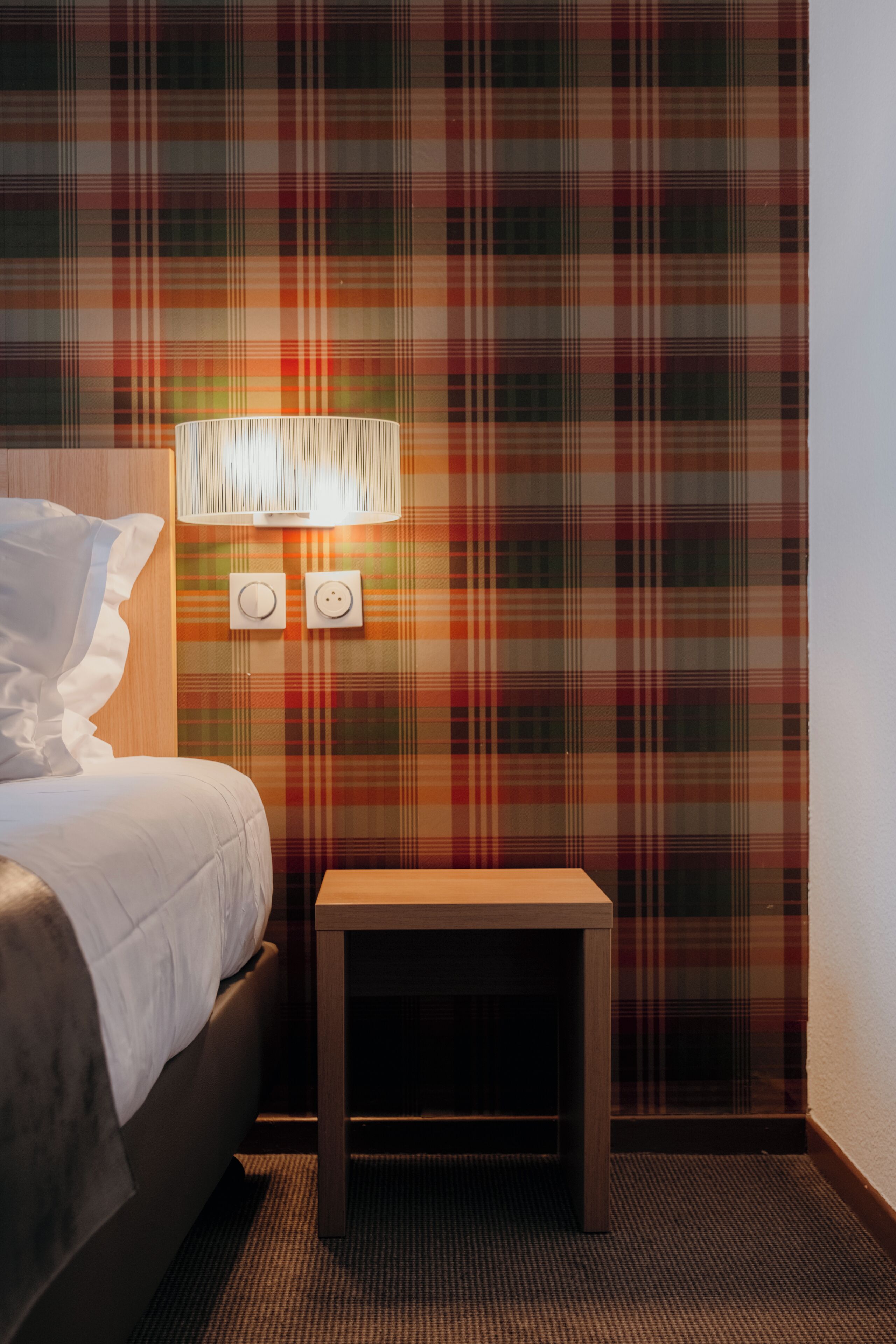 Une chambre d’hôtel avec un mur à motifs écossais, une lampe de chevet, une table de chevet et un lit avec une literie blanche.