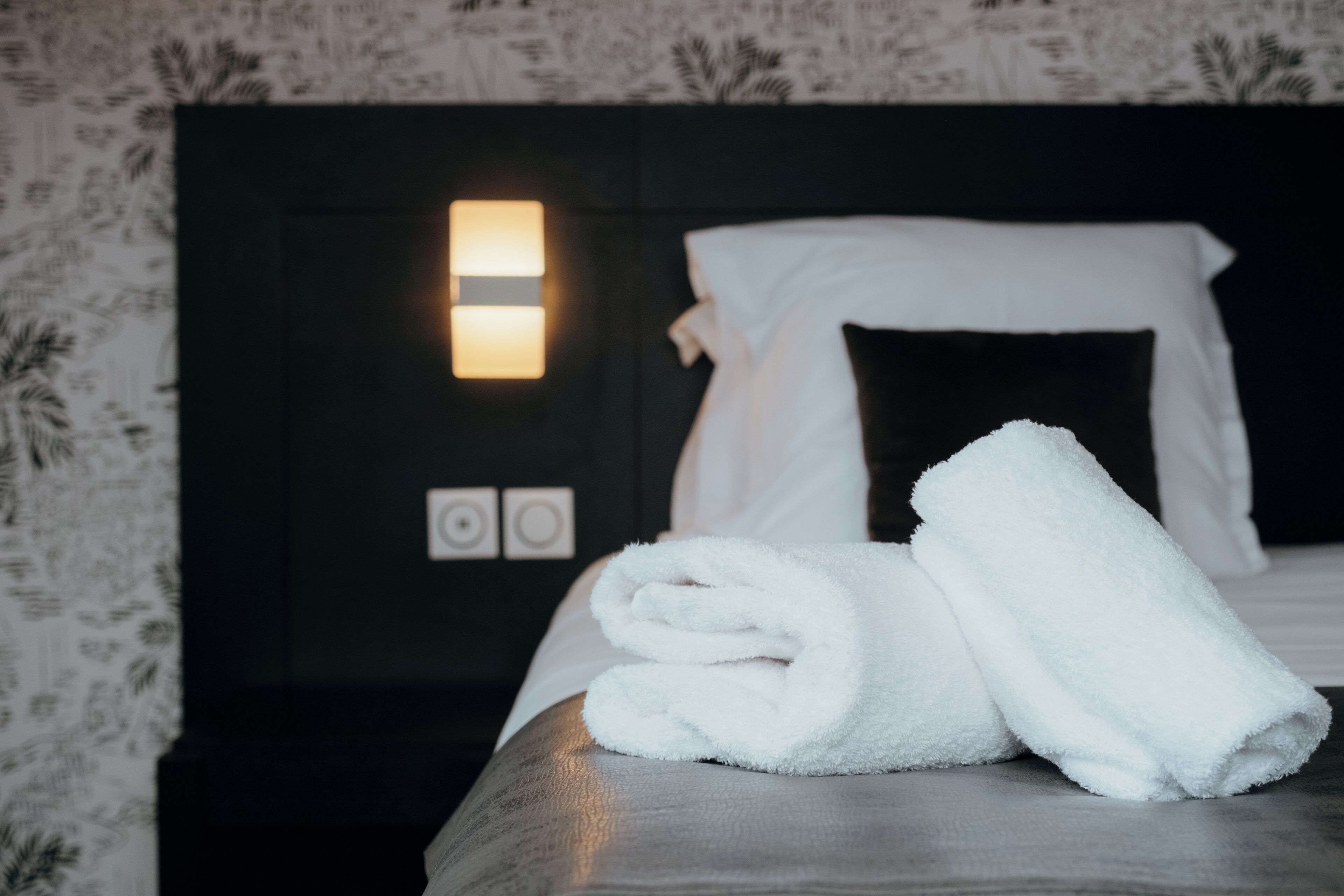 Una cama bien hecha con sábanas blancas y una toalla doblada, una lámpara de noche y una pared con estampado floral.