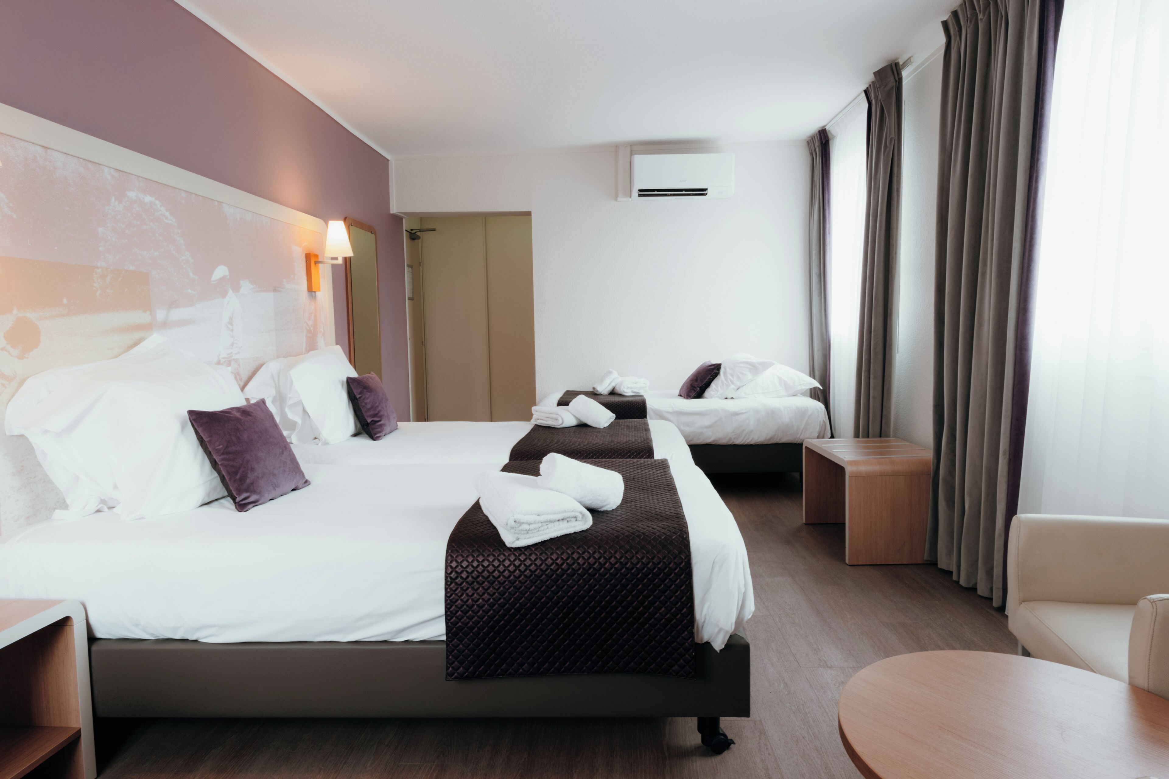 A hotel room with three beds, a sofa, a round table, and a wall-mounted air conditioner.