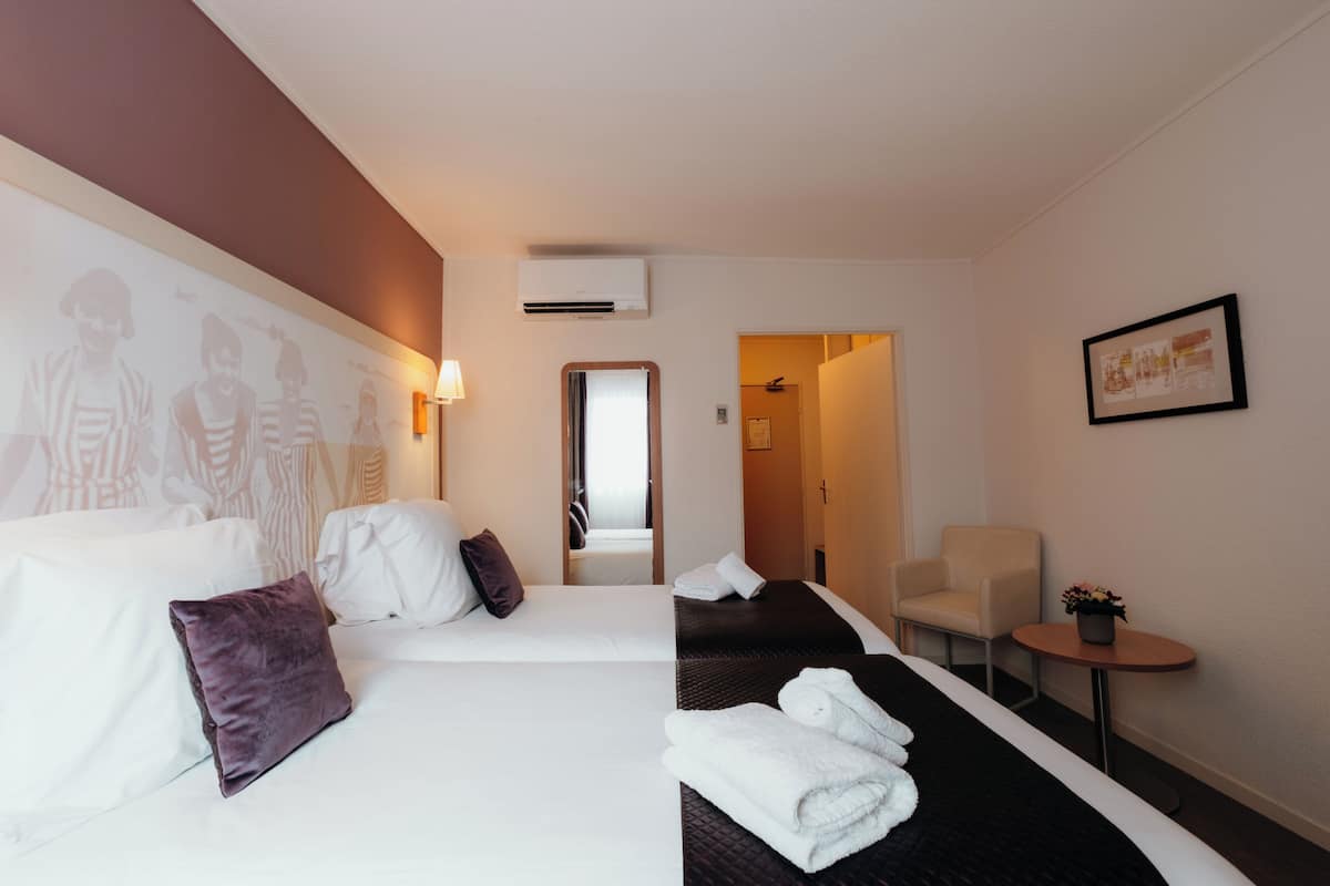 A hotel room with two beds, a small table with a plant, a chair, and a wall with a framed picture.