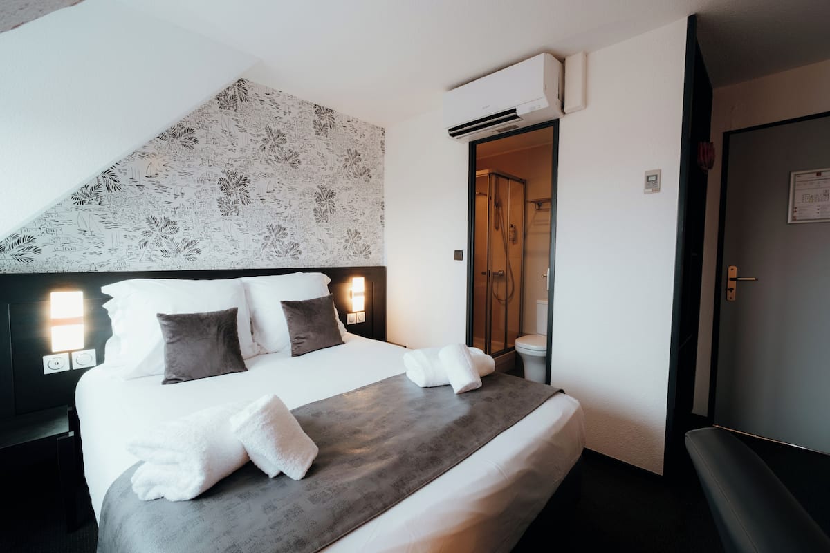 A hotel room with a large bed, a headboard with a floral pattern, a bedside table with a lamp, and an air conditioning unit.