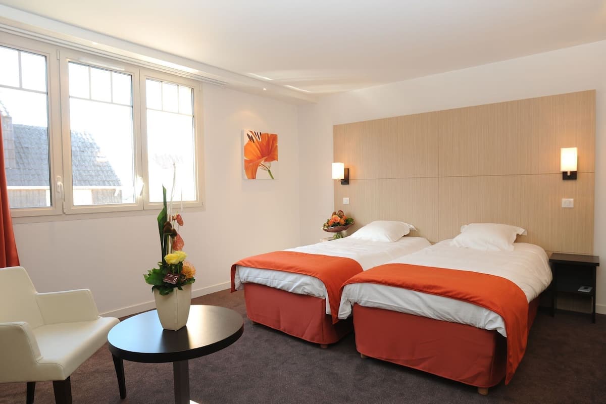 A hotel room with two beds, a small table, a chair, a vase with flowers, and a window with curtains.