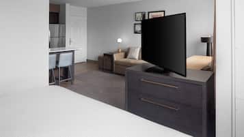 A modern hotel room with a flat-screen TV, a sofa, a desk with chairs, and a kitchenette.