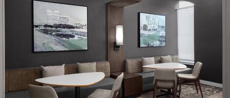 A modern hotel room with a seating area featuring a sofa, two round tables, and chairs. There are two large framed pictures on the wall, and a light fixture above the seating area.