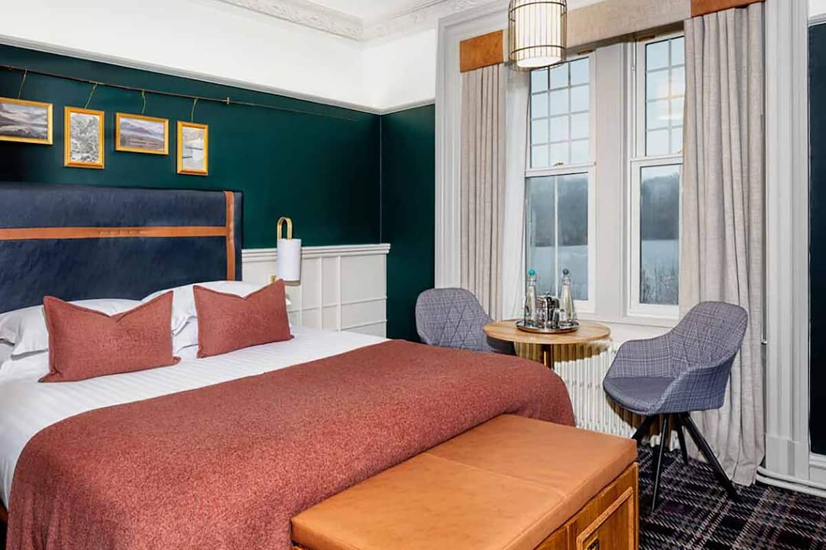 Une chambre d’hôtel avec un grand lit, un banc en bois, deux chaises, une petite table et une fenêtre avec des rideaux.
