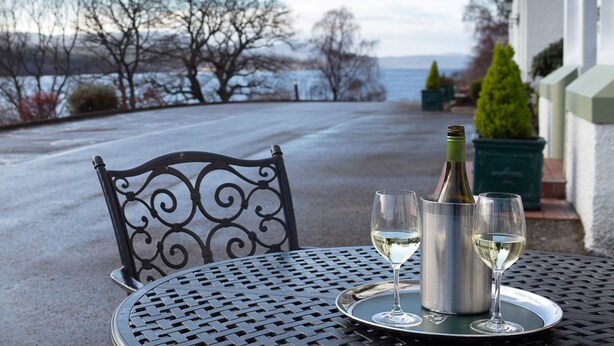 A table with a bottle and two glasses of white wine, a metal chair, and a view of trees and a building. - Loch Rannoch Hotel and Spa (Pitlochry)