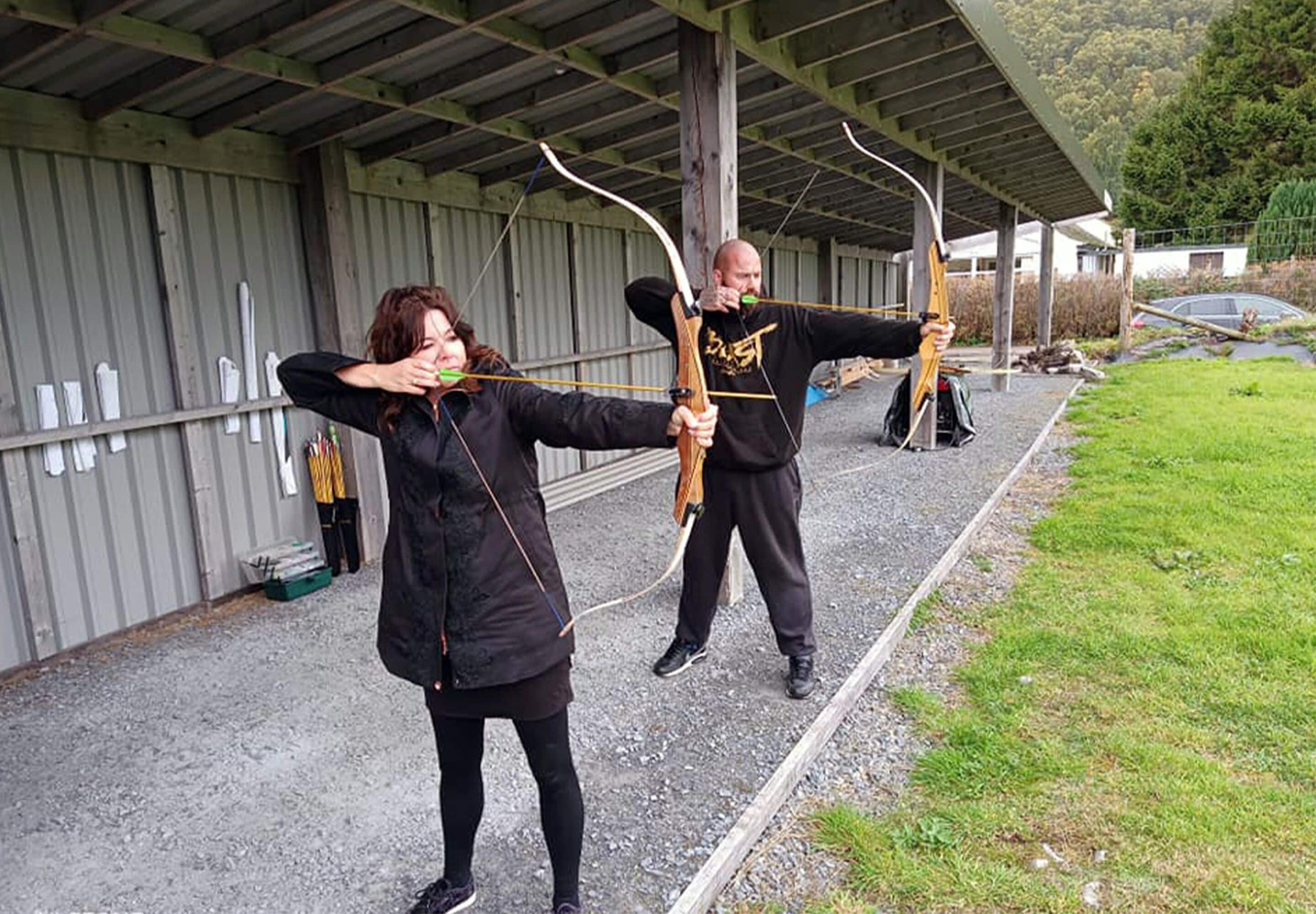 Two people practicing archery outdoors with a building and trees in the background.