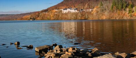 A lakeside view with a large building on a hill, surrounded by autumn trees and rocky shoreline.