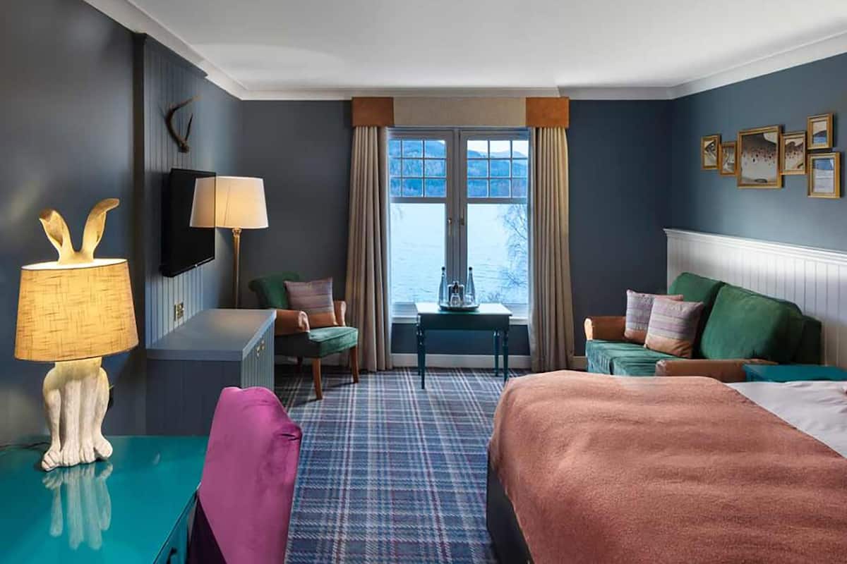 Une chambre d’hôtel avec un grand lit, un bureau avec une lampe, une chaise, une télévision et une fenêtre avec des rideaux.