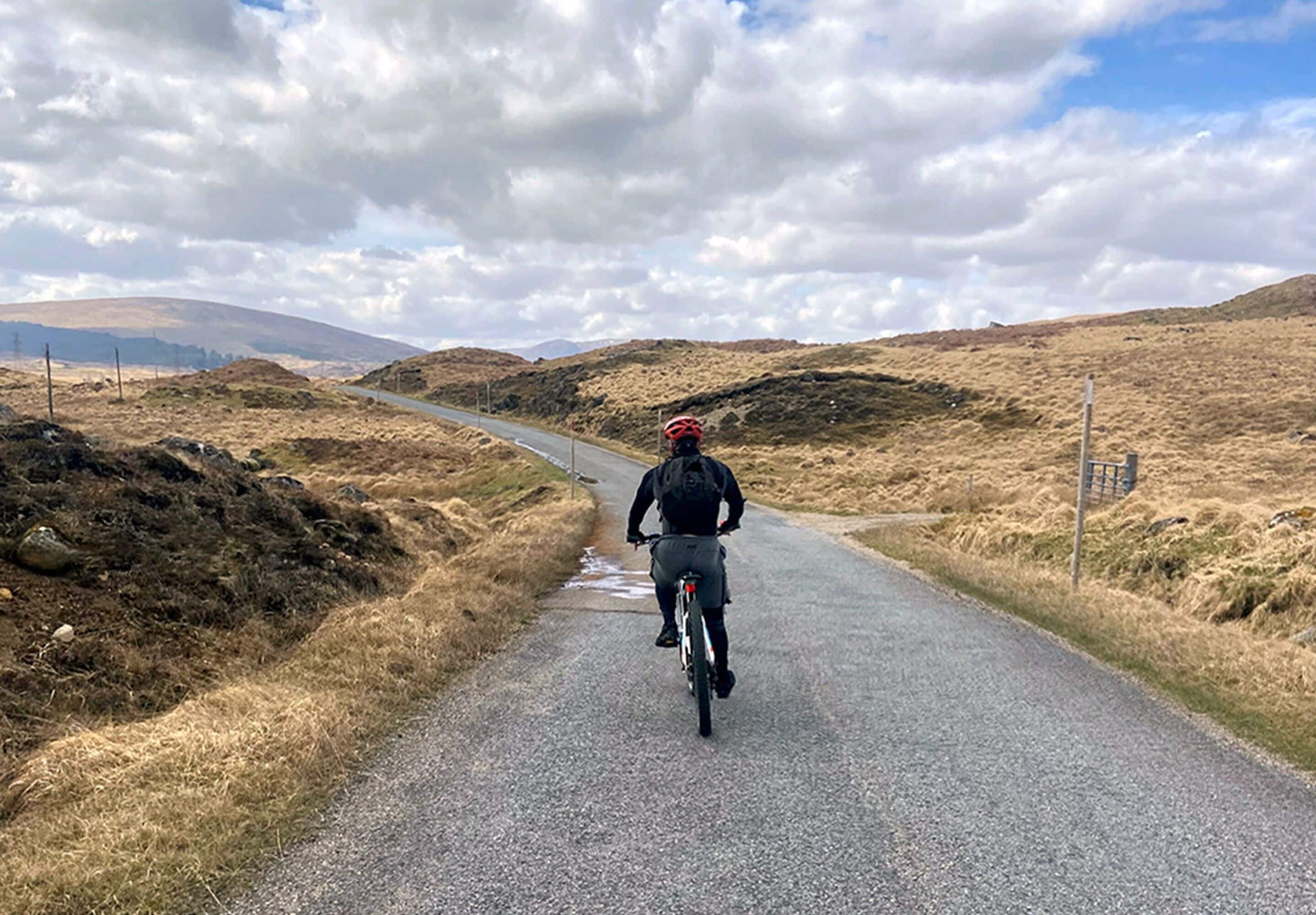 A cyclist on a gravel road with dry grass on either side and a cloudy sky.