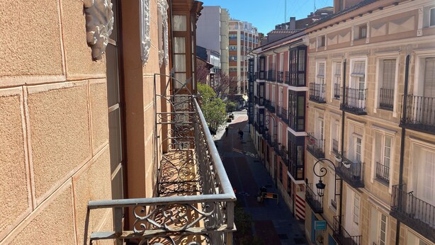 Un carrer urbà estret amb arquitectura europea tradicional, balcons amb baranes de ferro forjat i un cel blau clar.