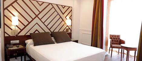 Habitación de hotel con una cama grande, dos lámparas de noche, un cabecero de madera, una mesita con una silla y vistas a través de una puerta abierta.