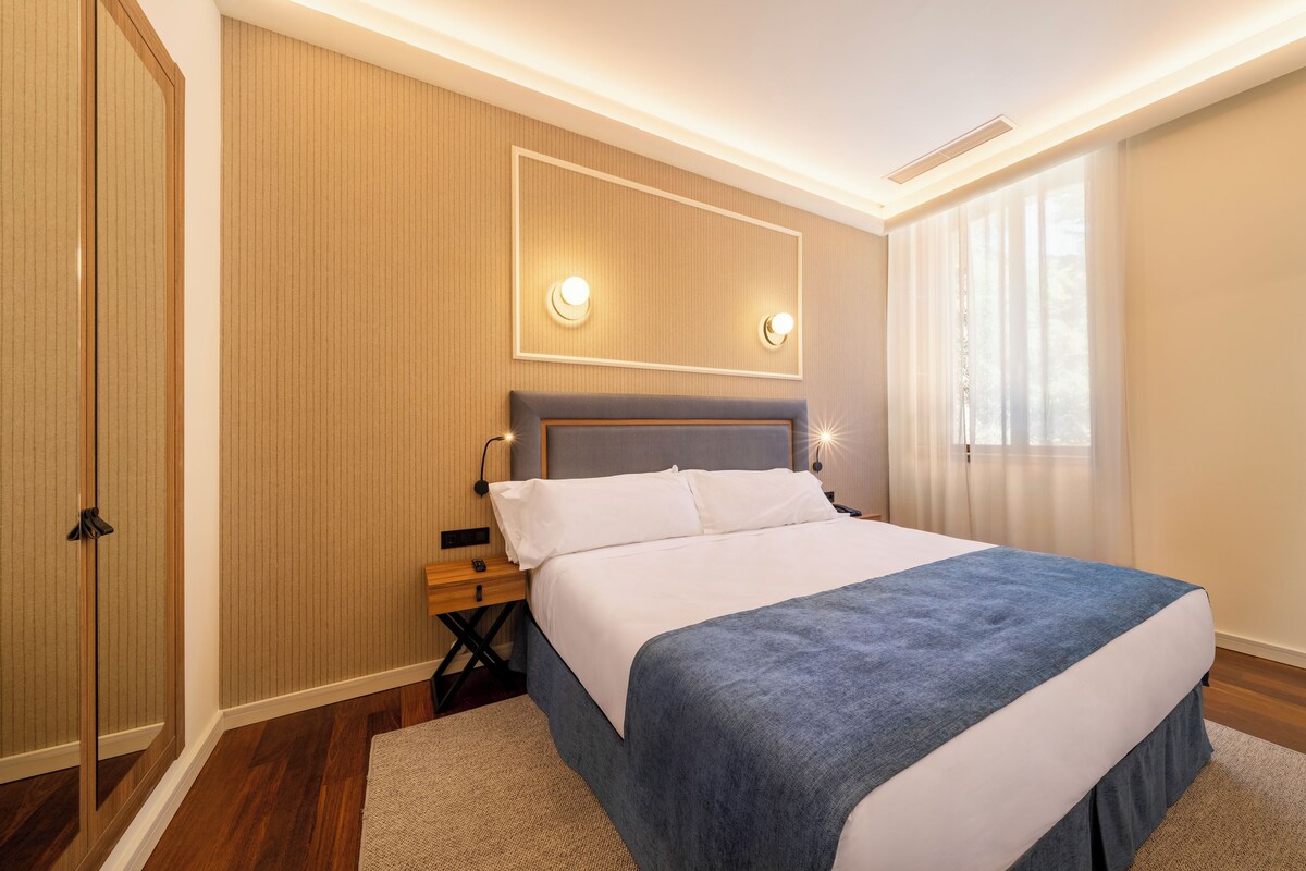 A modern hotel room with a large bed, wooden panel walls, a mirror, and a window with curtains.