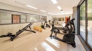 Une salle de sport moderne équipée de divers appareils d’exercice, d’un banc en bois et de grandes fenêtres qui laissent entrer la lumière naturelle et offrent une vue dégagée.