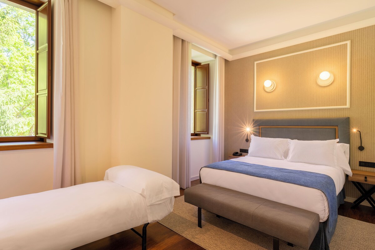 A hotel room with a large bed, a smaller bed, a bench, a nightstand, and a wall-mounted light fixture.