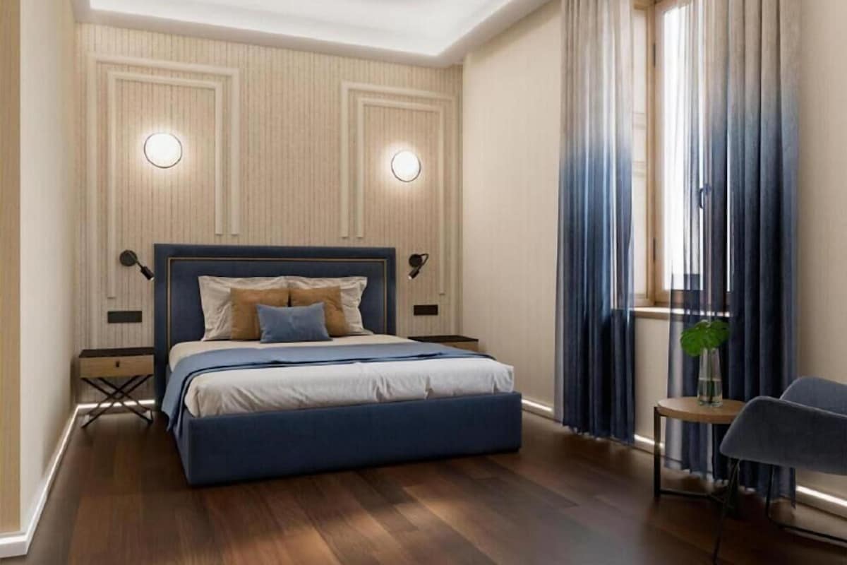 Ein Schlafzimmer mit einem großen Bett, zwei Nachttischlampen, einem kleinen Tisch mit Pflanze und einem Sessel.