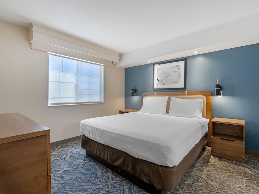 A hotel room with a bed, bedside tables, a desk, a window, and a wall-mounted picture.