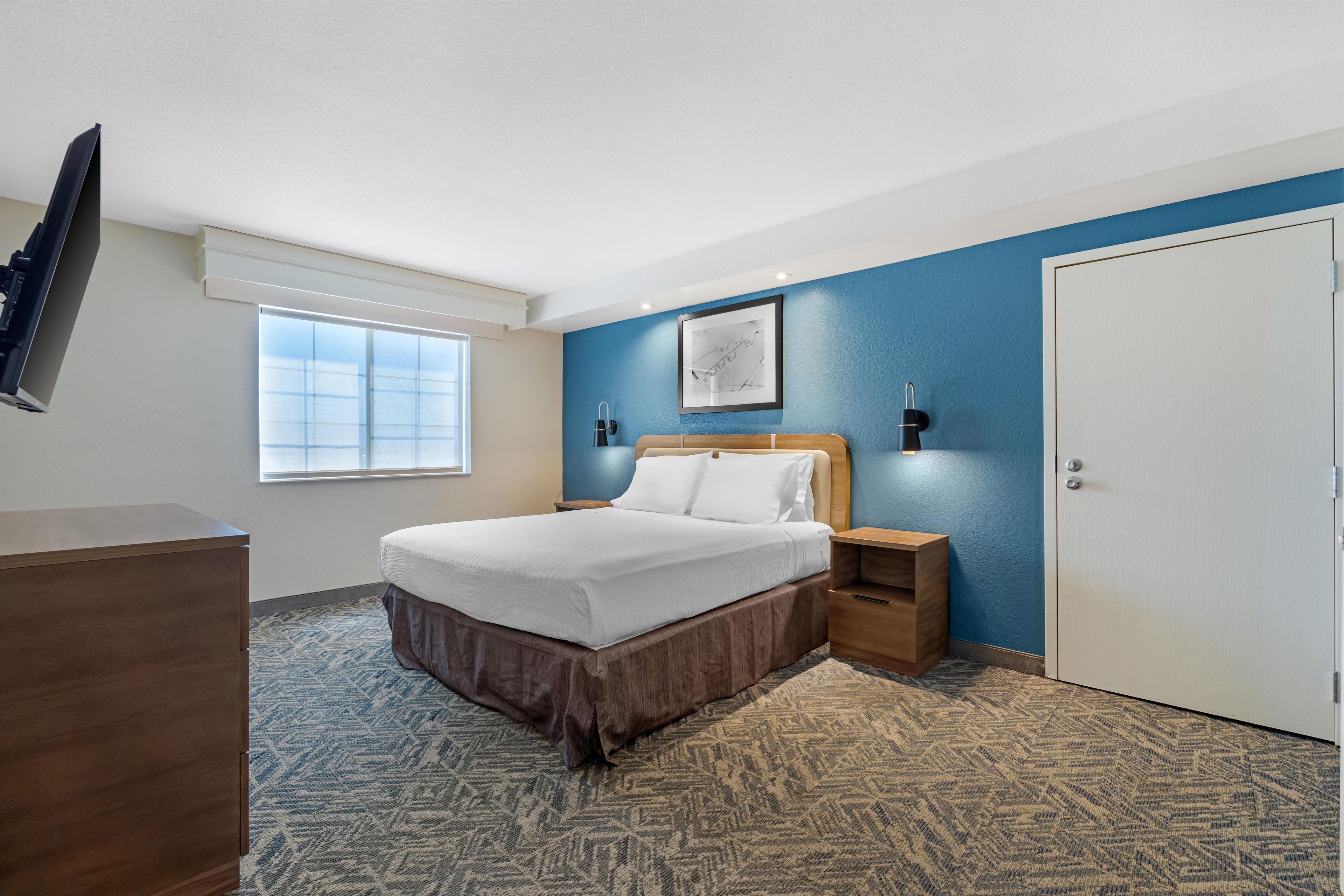 A hotel room with a bed, a nightstand, a TV, a window, and a door.