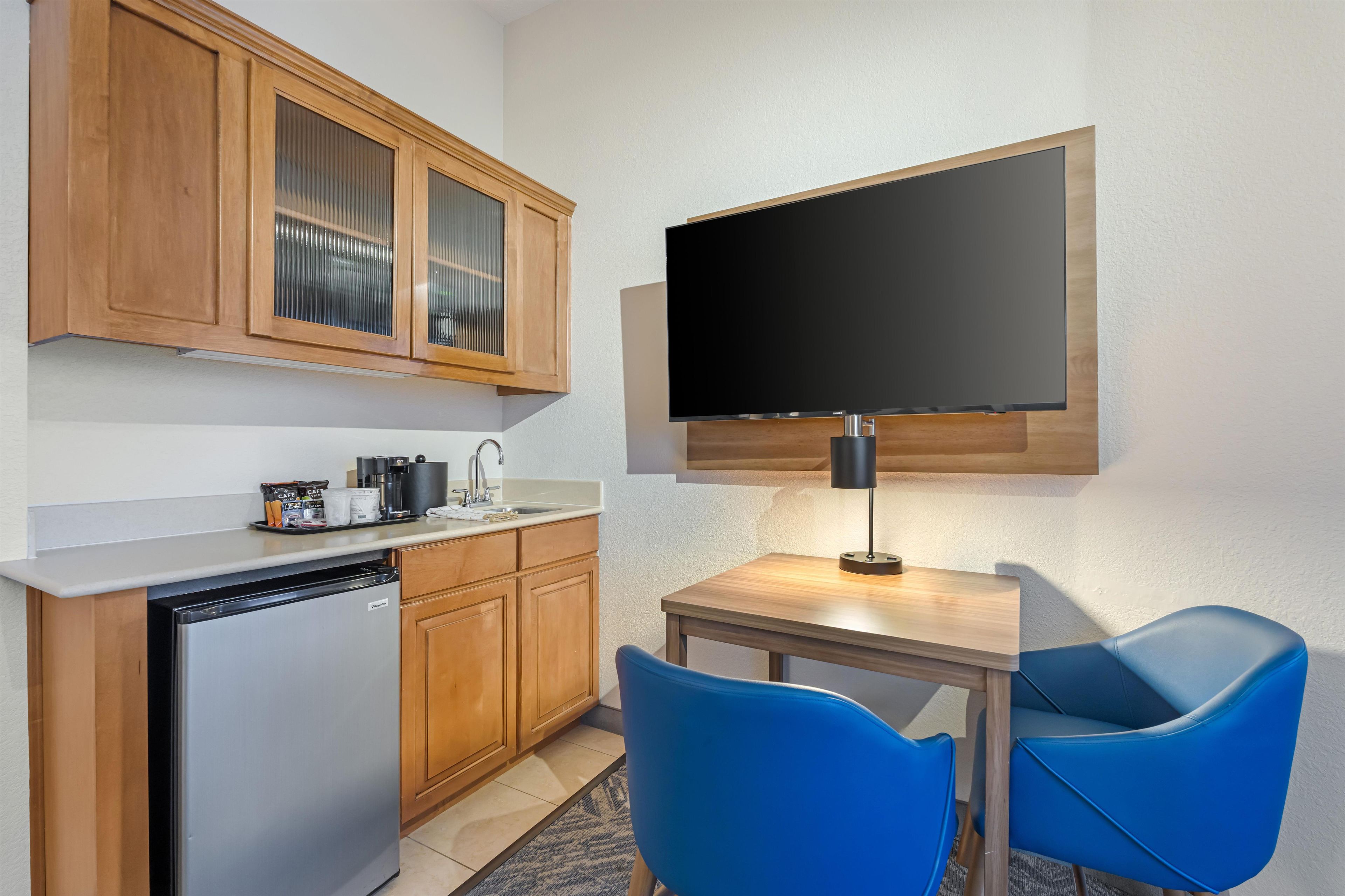 Uma cozinha compacta equipada com geladeira, máquina de café e bule. Há uma TV de tela plana fixada na parede acima de uma mesa de madeira com duas cadeiras azuis.