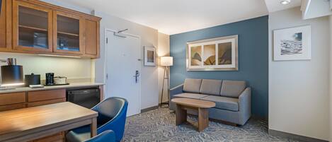 A compact hotel room with a kitchenette, a sofa, a dining table, and a patterned carpet.