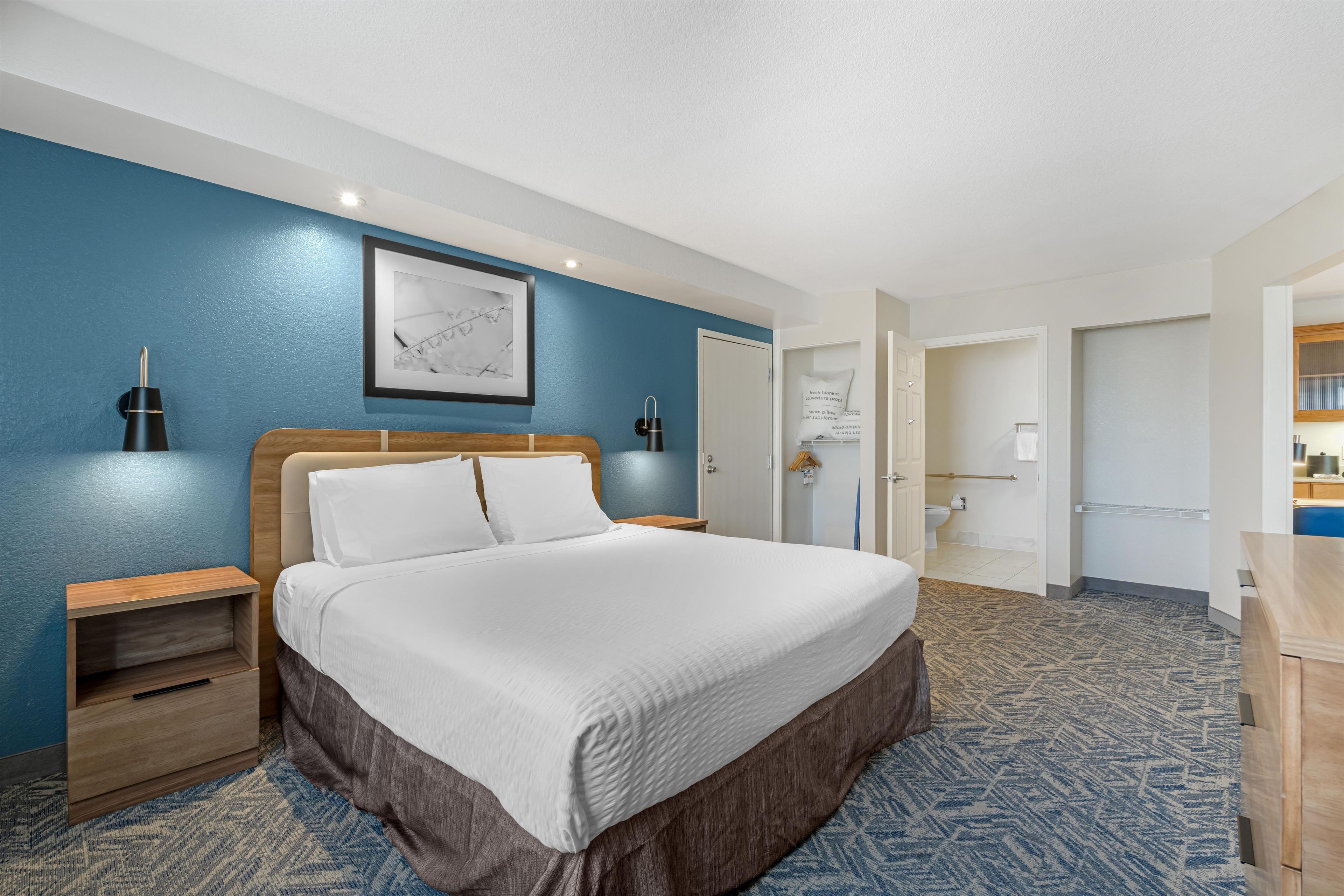 Ein Hotelzimmer mit einem großen Bett, einem Nachttisch, einer Wandleuchte, einem gerahmten Bild und einem durch eine offene Tür einsehbaren Badezimmer.