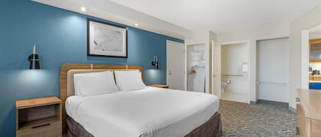 Habitación de hotel con una cama grande, una mesita de noche, una lámpara de pared, un cuadro enmarcado y un baño visible a través de una puerta abierta.