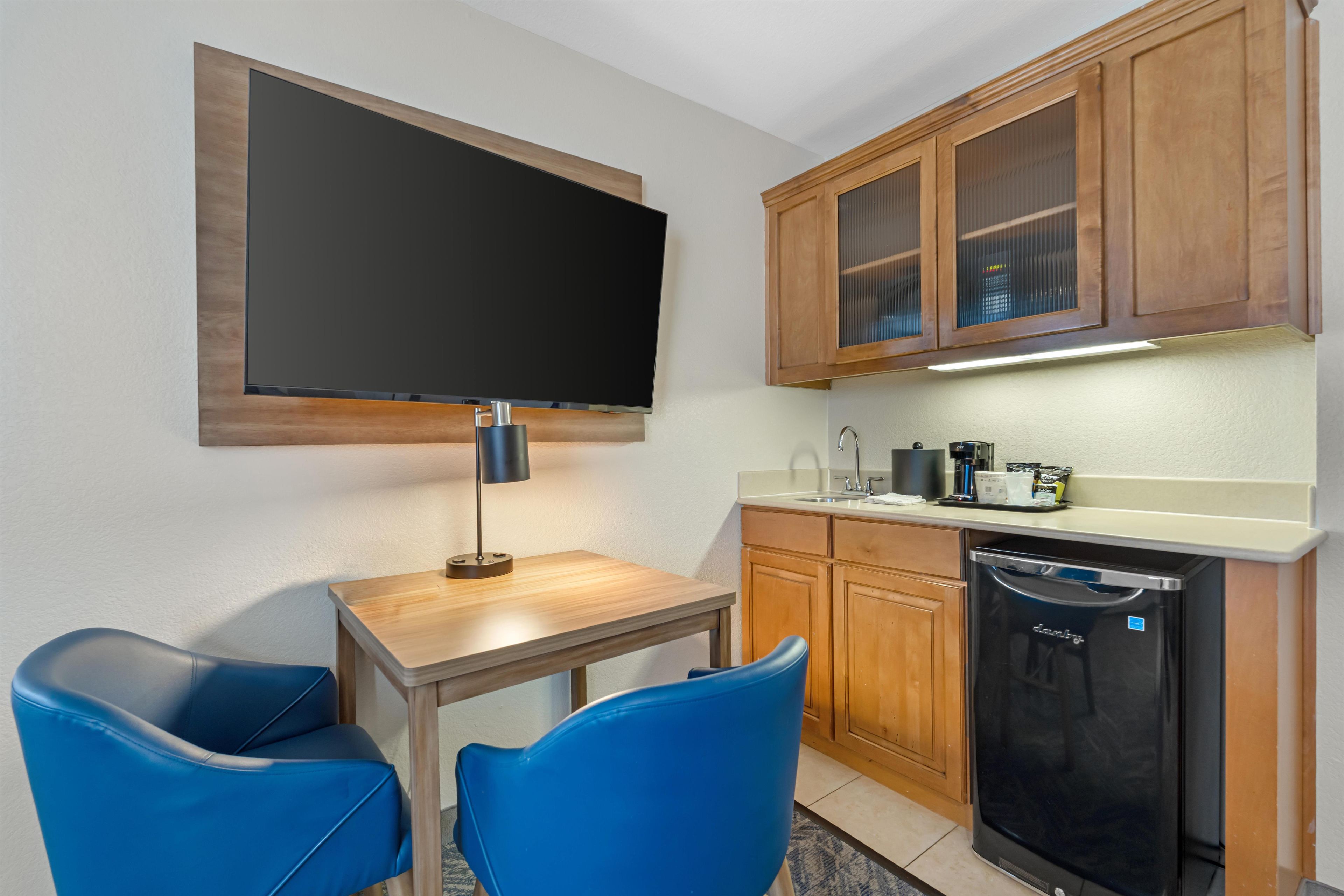 A compact kitchen with a built-in dishwasher, a flat-screen TV mounted above a wooden table, and two blue chairs.