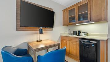 A compact kitchen with a built-in dishwasher, a flat-screen TV mounted above a wooden table, and two blue chairs.