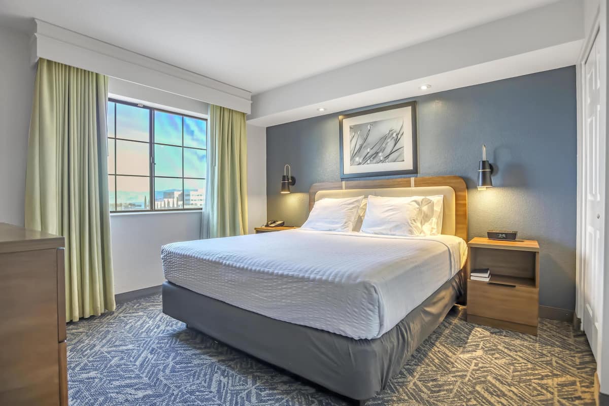 A hotel room with a large bed, a nightstand, a wall-mounted lamp, and a window with curtains.