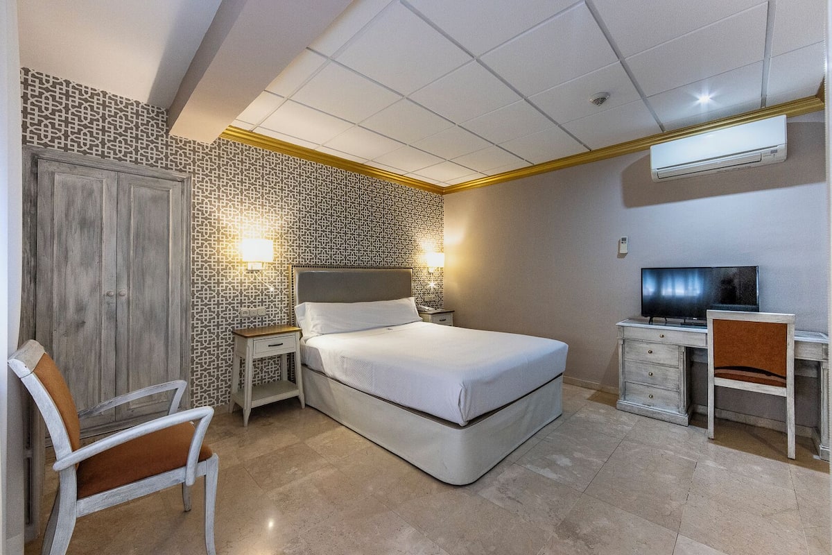 A bedroom with a bed, bedside table, lamp, TV, desk, chair, and an air conditioning unit.