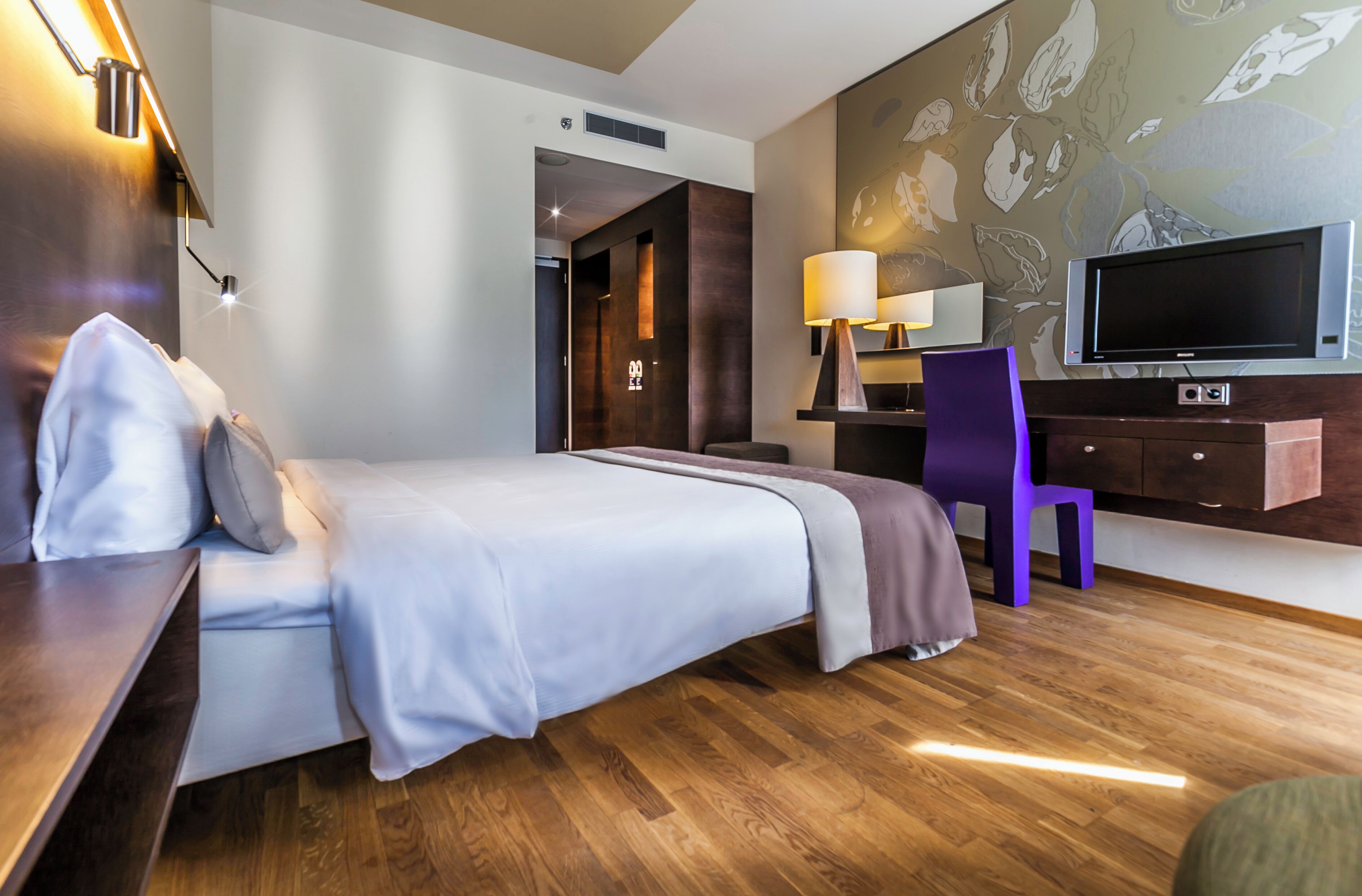 Een moderne hotelkamer met een bed, nachtkastje, lamp, televisie en een paarse stoel.