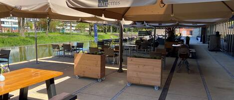 An outdoor café area with tables and chairs under large umbrellas, a wooden planter, and a bench.