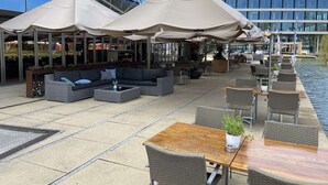 An outdoor seating area with wicker furniture, umbrellas, and a view of a modern building. - Hotel Artemis Amsterdam (Amsterdam)