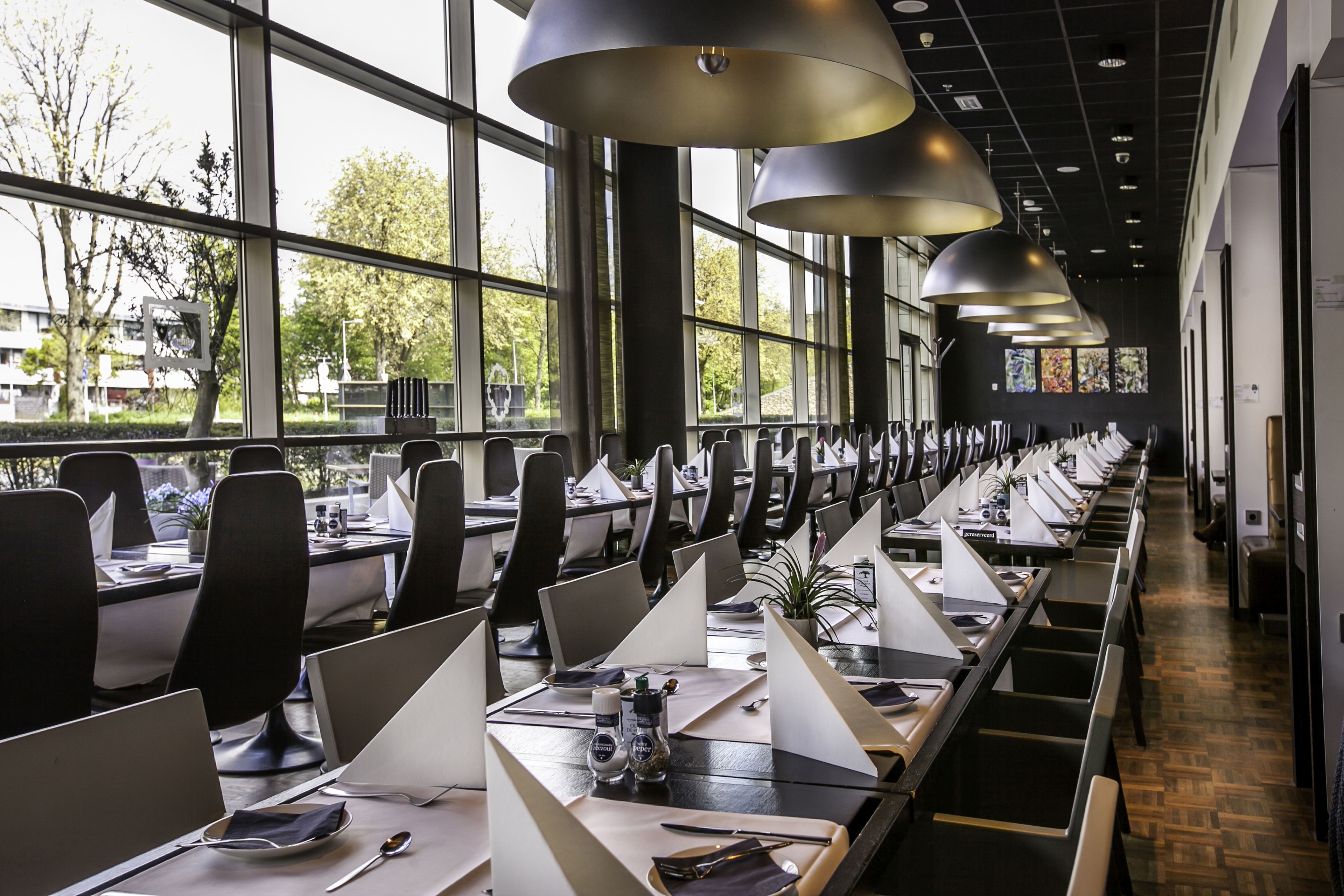 A modern office space with long tables, individual workstations, and large windows offering views of the outdoors.