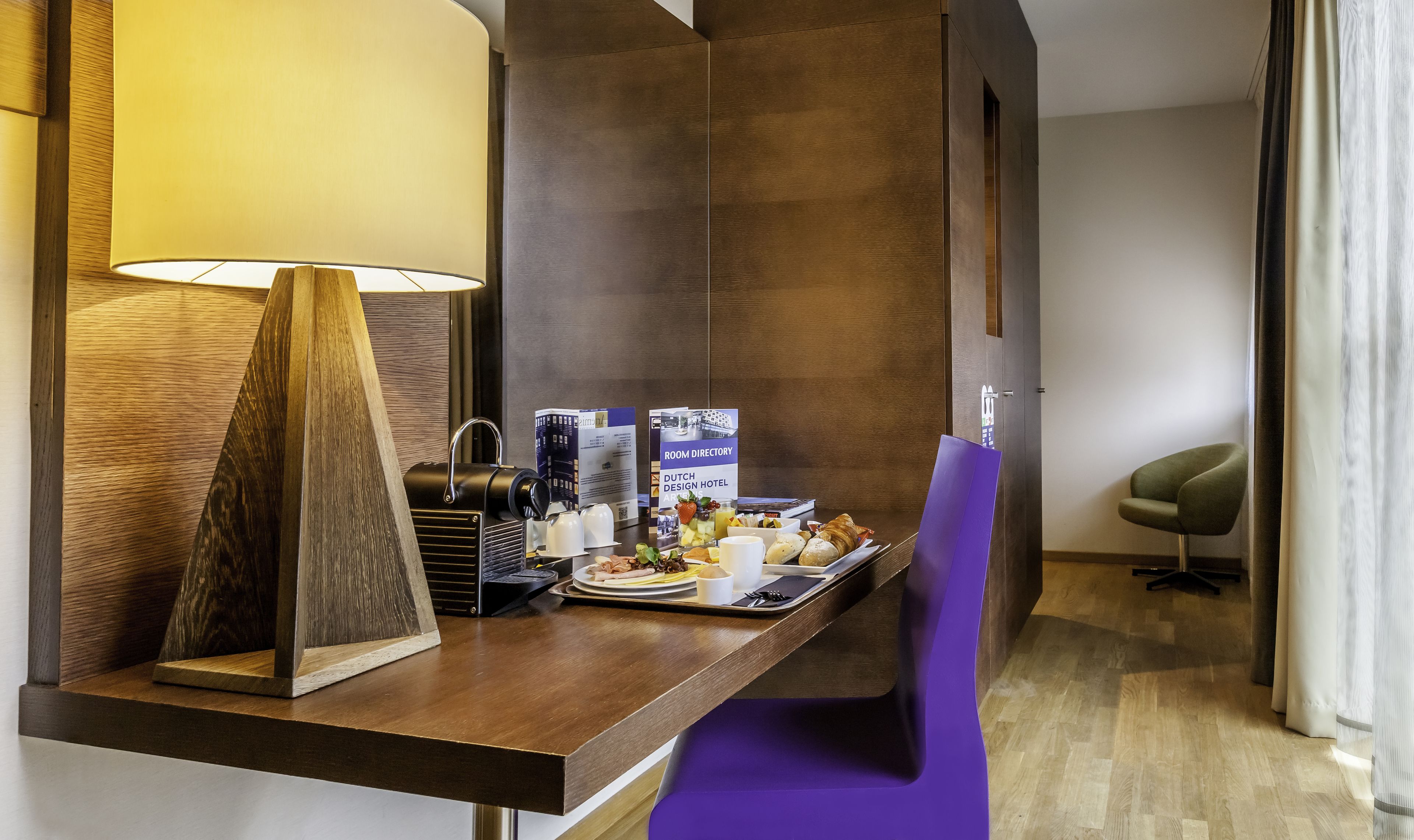 A hotel reception area with a wooden desk, a lamp, a room directory, and a purple chair.