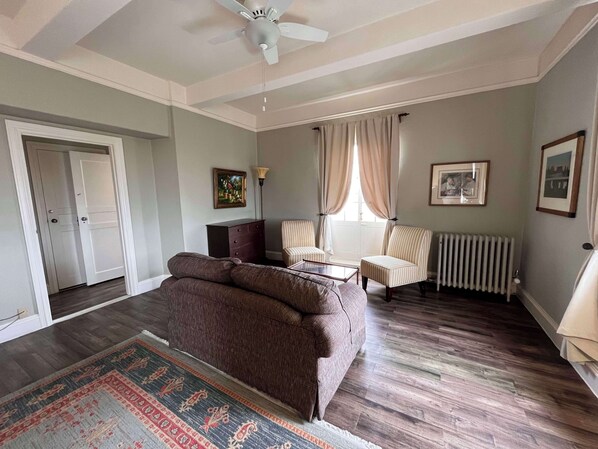 A living room with a brown sofa, two striped armchairs, a coffee table, and a patterned rug. There is a ceiling fan, a radiator, and two framed pictures on the walls. - Berkeley City Club (Berkeley)