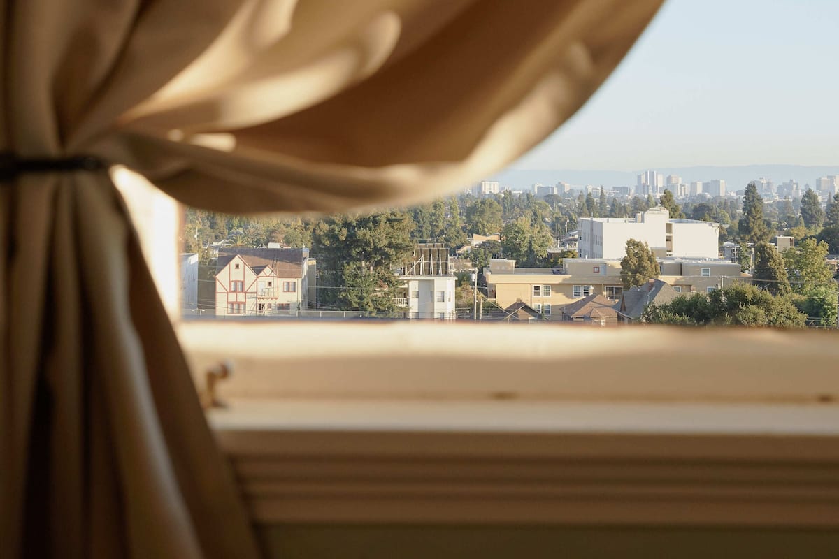 Blick durch ein Fenster mit Vorhängen auf eine Stadtlandschaft mit Gebäuden und Bäumen.