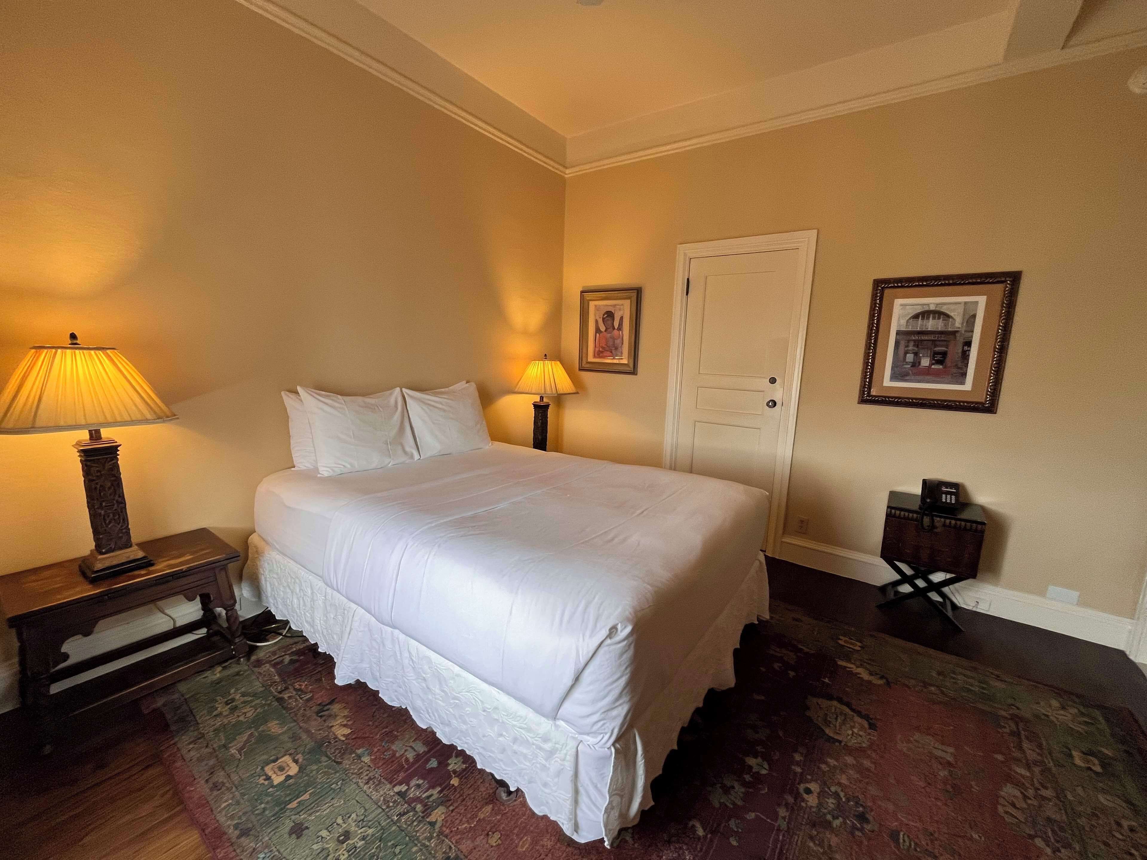 A neatly made bed with white linens in a hotel room. There is a wooden nightstand with a lamp and a small table with a telephone. The room has a carpeted floor and a door with a picture frame above it.