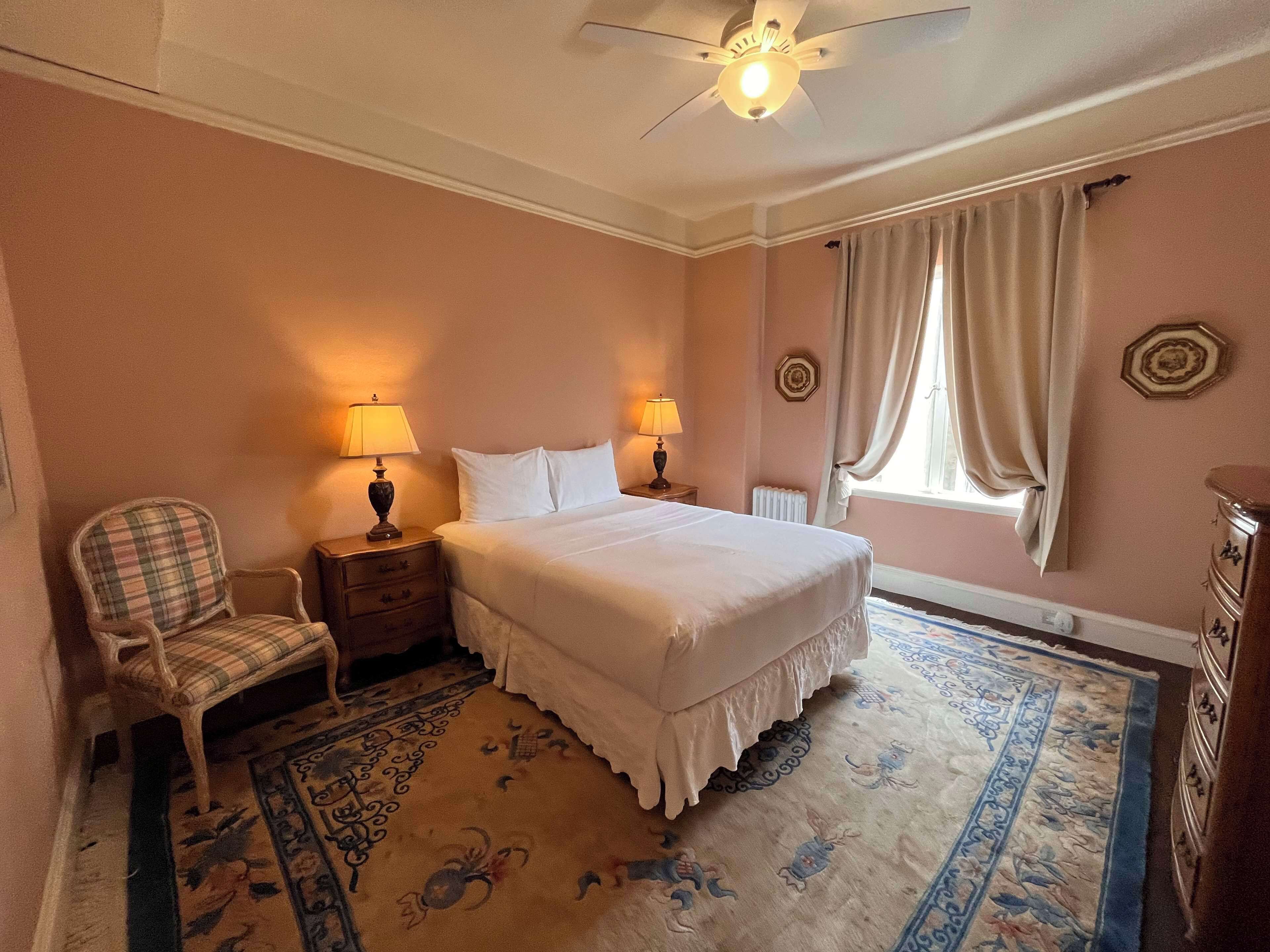 A bedroom with a bed, two bedside tables with lamps, a chair, a radiator, a window with curtains, and a patterned rug.