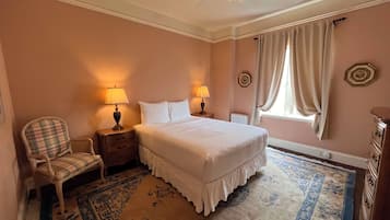Une chambre à coucher avec un lit, deux tables de chevet avec des lampes, une chaise, un radiateur, une fenêtre avec des rideaux et un tapis à motifs.