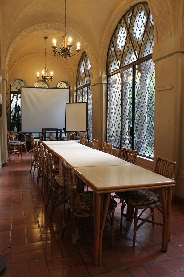 Un salón de conferencias con una mesa larga, sillas de mimbre, una pantalla de proyector y grandes ventanales de arco con celosías metálicas.