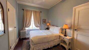 A bedroom with a bed, two bedside lamps, a window with curtains, a radiator, and a patterned rug.