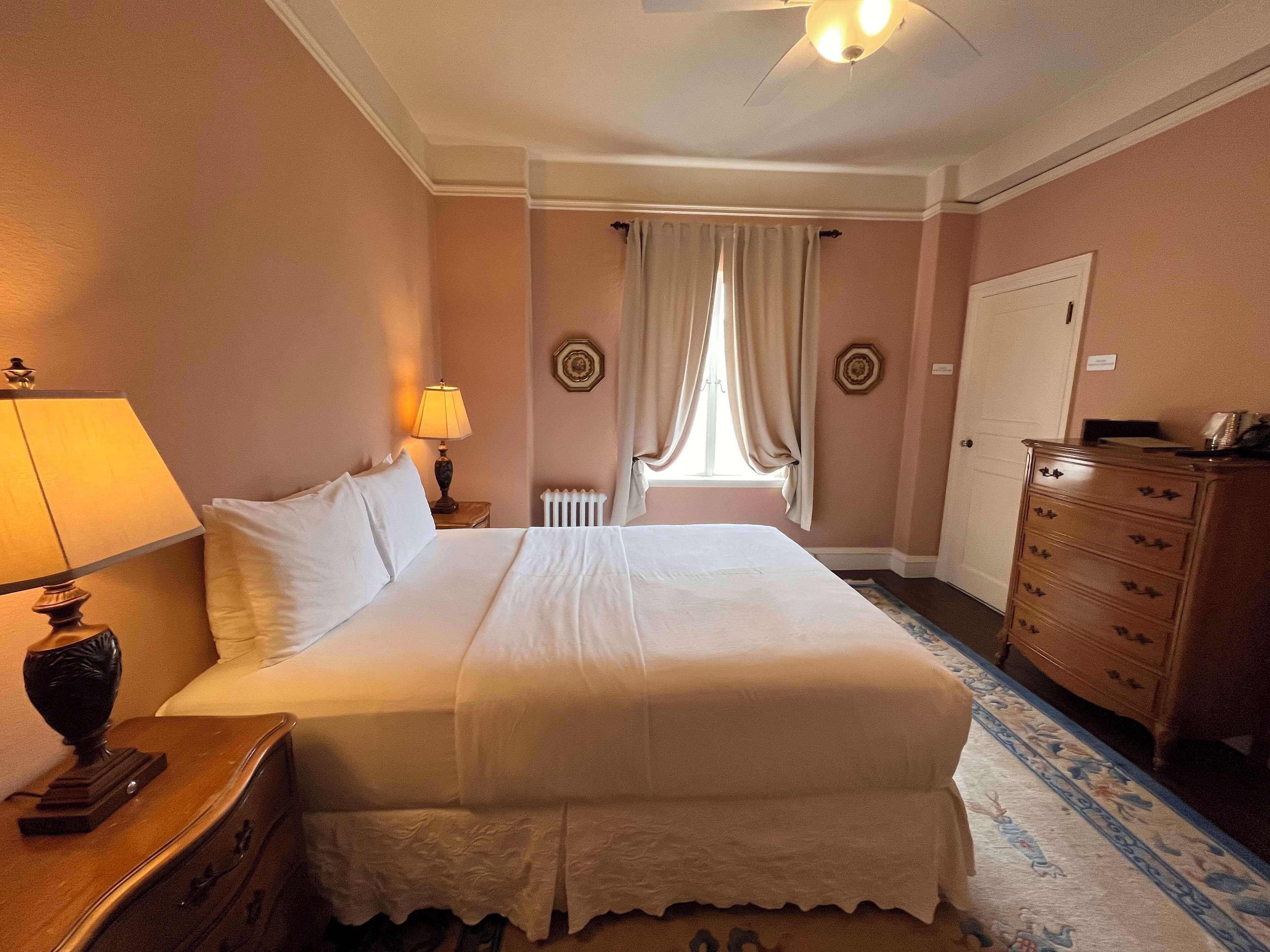 Habitación de hotel con una cama grande, un aparador, una lámpara y un ventanal con cortinas.